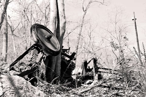 The rusted wreck of an overturned vehicle lying among bare trees and rocks in stark black-and-white woodland.