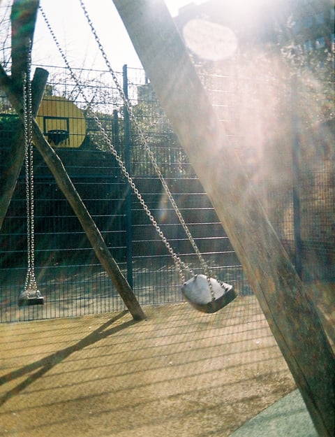 Empty chain swings in a fenced playground, lit by warm low sun with bright lens flare.