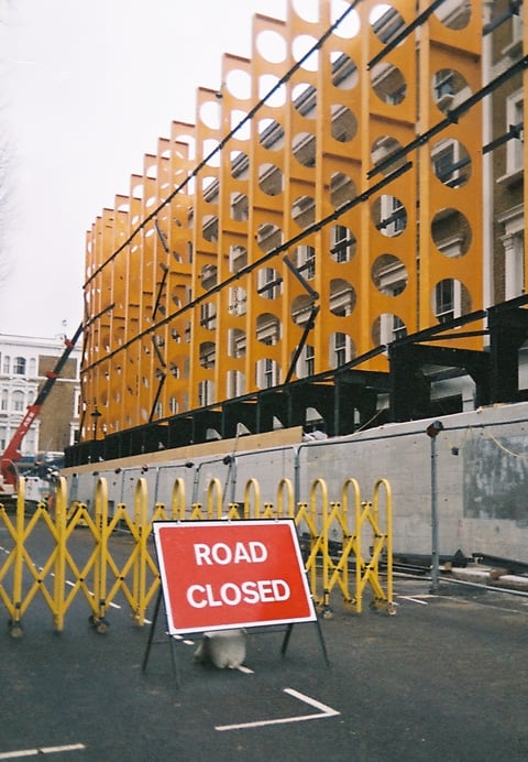 A red road-closed sign and yellow barriers before a bright orange perforated building facade under grey sky.