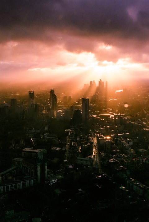 Aerial view of a hazy city skyline at sunset, with golden light rays piercing through dark clouds.