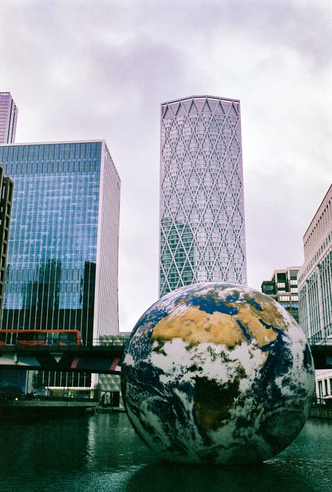 A giant floating globe sculpture of Earth rests on water among glass office towers, with a light railway train crossing a bridge under a grey sky.