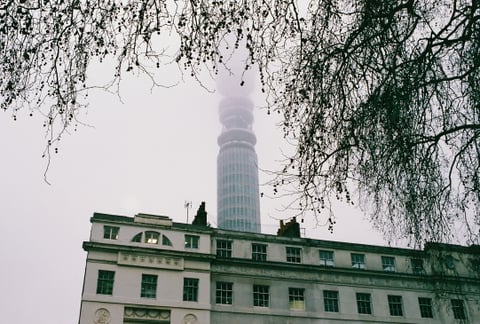 The BT Tower fades into mist above a pale building, framed by bare branches under grey sky.