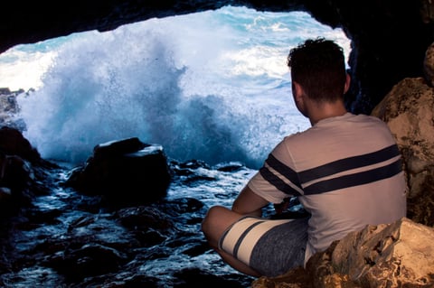 A person sitting near dark rocks while waves crash into a sea cave.