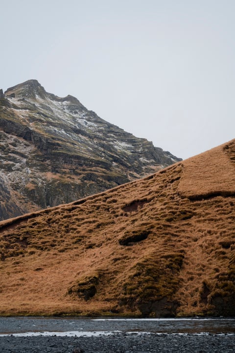 A steep slope of rust-coloured grass below a snow-dusted rocky peak, with a dark gravel riverbed in the foreground.
