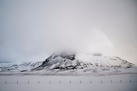 A snow-covered mountain with its summit lost in cloud, a fence line crossing the white plain below.
