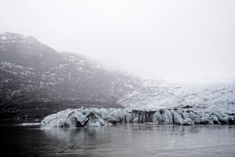 A glacier tongue meeting a still, icy lagoon beneath misty dark mountains.