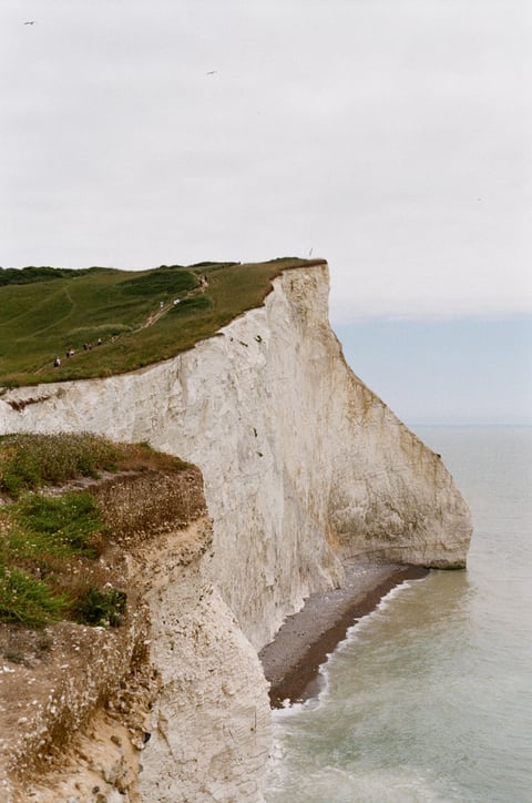 White chalk cliffs topped with green grass descend to a pebble beach beside a pale grey sea.