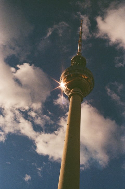 The Berlin TV Tower seen from below, its sphere catching a sunburst against blue sky and clouds.