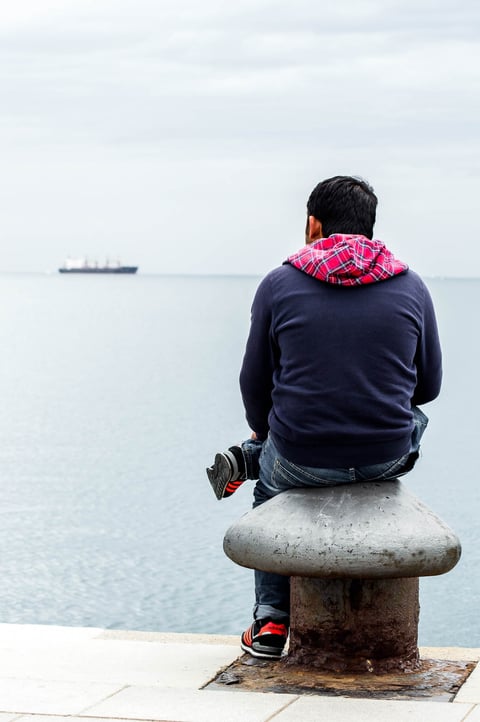 A person in a dark jacket sitting on a bench and looking over water.